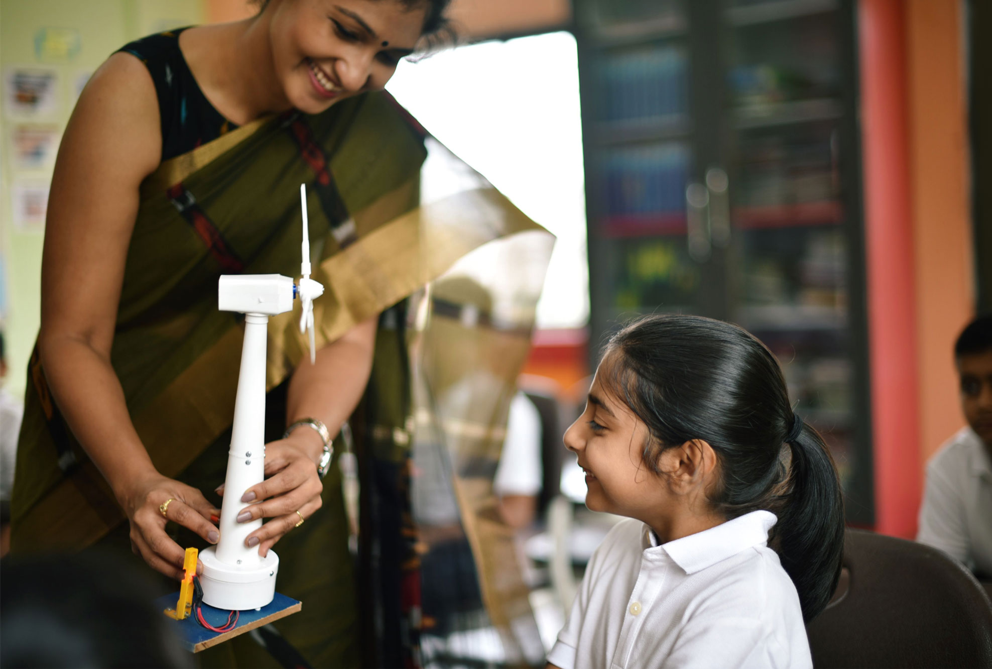 A teacher helping a pupil with a wind turbine model in a school classroom.