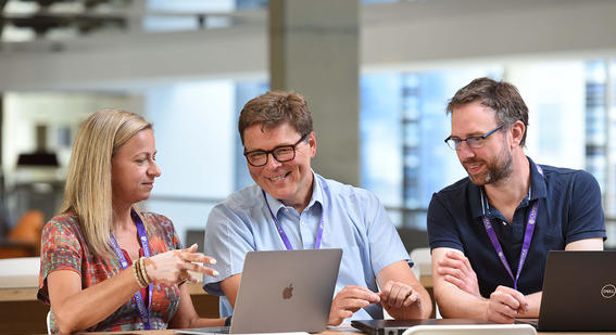 Three Cambridge colleagues sit in front of their laptops smiling while the female colleague points to her screen for her other two male colleagues to look at