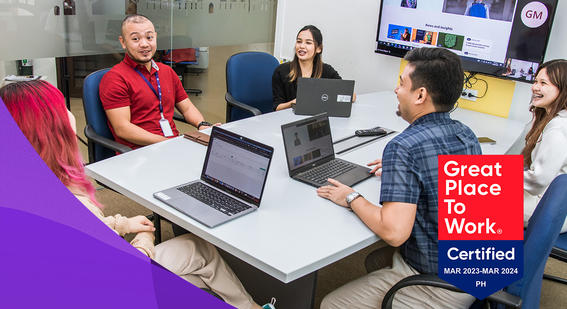 Manila colleagues sit in chairs around a desk during a meeting discussing work in front of laptops smiling with GPTW certification logo at bottom right