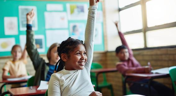 a young student holds her hands up in class while sat behind a desk